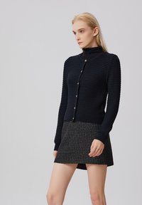 Gilet en tricot bleu marine avec un design texturé, doté d'un col haut, de manches longues et de boutons dorés. Assorti avec une jupe à motifs.