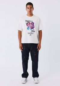 Camiseta blanca con un gráfico floral en rosa y azul, combinada con jeans de mezclilla oscuros decorados con costuras en forma de estrella y zapatillas blancas.