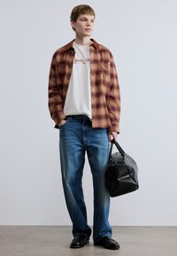 Κόκκινο και καφέ πουκάμισο καρό πάνω από λευκό μπλουζάκι, μπλε τζιν και μαύρα παπούτσια. Κρατώντας μία μαύρη τσάντα με υφή. Ουδέτερο φόντο.