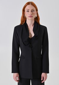 Blazer nero con colletto a scialle e chiusura con un bottone. Presenta due tasche frontali e una vestibilità su misura. Tessuto liscio e design strutturato.