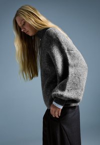 Szürke, túlméretezett kötött pulóver magas nyakkal és texturált anyaggal, sötét nadrággal párosítva. A háttér lágy kék.