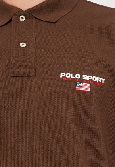 Camisa polo castanha com um tecido texturizado, apresentando colarinho e dois botões. O logótipo bordado diz "POLO SPORT" em branco e vermelho, com uma pequena bandeira americana.