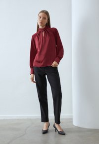 Burgundy long-sleeve blouse with a tie-neck detail, paired with black slim-fit trousers and black pointed-toe heels. Smooth fabric textures.