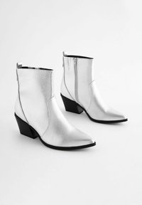 Botas de vaquero metálicas plateadas hasta el tobillo con una superficie texturizada, punta afilada y tacón cuadrado. Incluyen una cremallera lateral y un lazo en la parte posterior.