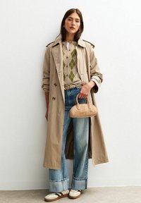 Donna con un trench beige, pullover a rombi, jeans risvoltati, cintura marrone, con in mano una piccola borsa beige, in piedi contro un muro bianco.