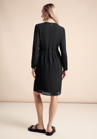 Robe noire plissée avec des manches longues, taille cintrée et un détail de bouton au dos. Associée à des sandales noires plates.