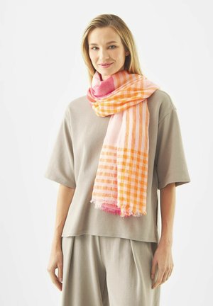 Vrouw die een beige losse top en broek draagt, met een roze-oranje geruite sjaal om haar nek gewikkeld, die zachtjes glimlacht.