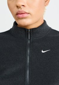 Czarna polarowa kurtka na zamek z wysokim kołnierzem i subtelnym logo Nike. Tekstura wydaje się miękka, z kontrastowym zamkiem i bez widocznych wzorów.