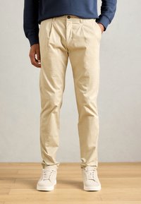 Beige cotton trousers with a relaxed fit, featuring a button closure and front pleats, paired with white athletic sneakers.