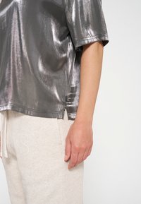 Metallic grauer Kurzarm-Top mit glatter Textur und leichtem Glanz, ausgestattet mit seitlichen Schlitzen und einem Etikett am Saum, kombiniert mit hellen Jogginghosen.