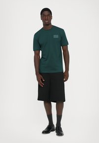 Tumši zaļa T-krekls ar īsām piedurknēm, apaļu kaklu un mazu kabatiņu. Valkāts kopā ar melnām šortiem un melnām loafer kurpēm, pieskaņots pelēkām zeķēm.