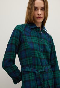 Vestido de cuadros en verde, azul y negro; cuenta con mangas largas, cierre de botones y cintura atada. Confeccionado con tela suave y texturizada.