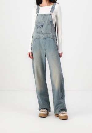 Femme portant une salopette en jean bleu délavé, un haut blanc à manches longues et des chaussures beiges doublées de fourrure, debout sur un fond blanc uni.