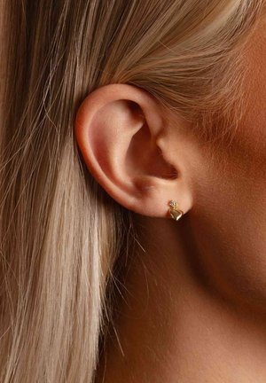 Ohrstecker in Herzform aus Gold mit kleinem Edelsteinakzent. Der Ohrring ist vor einem neutralen Ohr und glattem blondem Haar zu sehen.