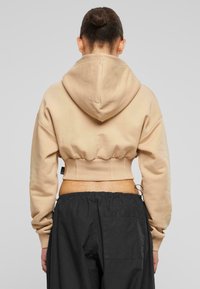 Felpa corta beige con cappuccio regolabile, vita elasticizzata aderente e polsini a costine. Abbinata a pantaloni neri larghi.