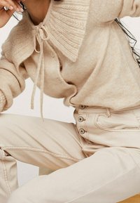 Pull en tricot beige avec un col large texturé et des liens, associé à un pantalon taille haute beige clair avec des accents de boutons.