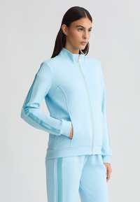 Giacca leggera azzurro chiaro con zip e colletto alto, caratterizzata da inserti in rete sulle maniche e tasche laterali, realizzata in un morbido tessuto testurizzato.