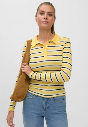 Jeune femme portant un polo à manches longues rayé jaune et un jean bleu, tenant un sac à bandoulière en daim marron, bras croisés.