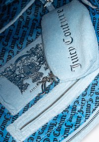 Ljusblå blixtlås-påse med texturerad tyg, med silverfärgad hårdvara, intrikata detaljer och varumärket "Juicy Couture" tydligt tryckt.