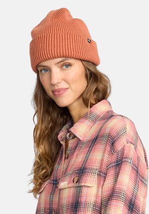 Berretto lavorato a maglia color ruggine con texture a coste, design aderente e una piccola etichetta nera. Abbinato a una camicia a quadri nei toni del rosa e dell'azzurro.
