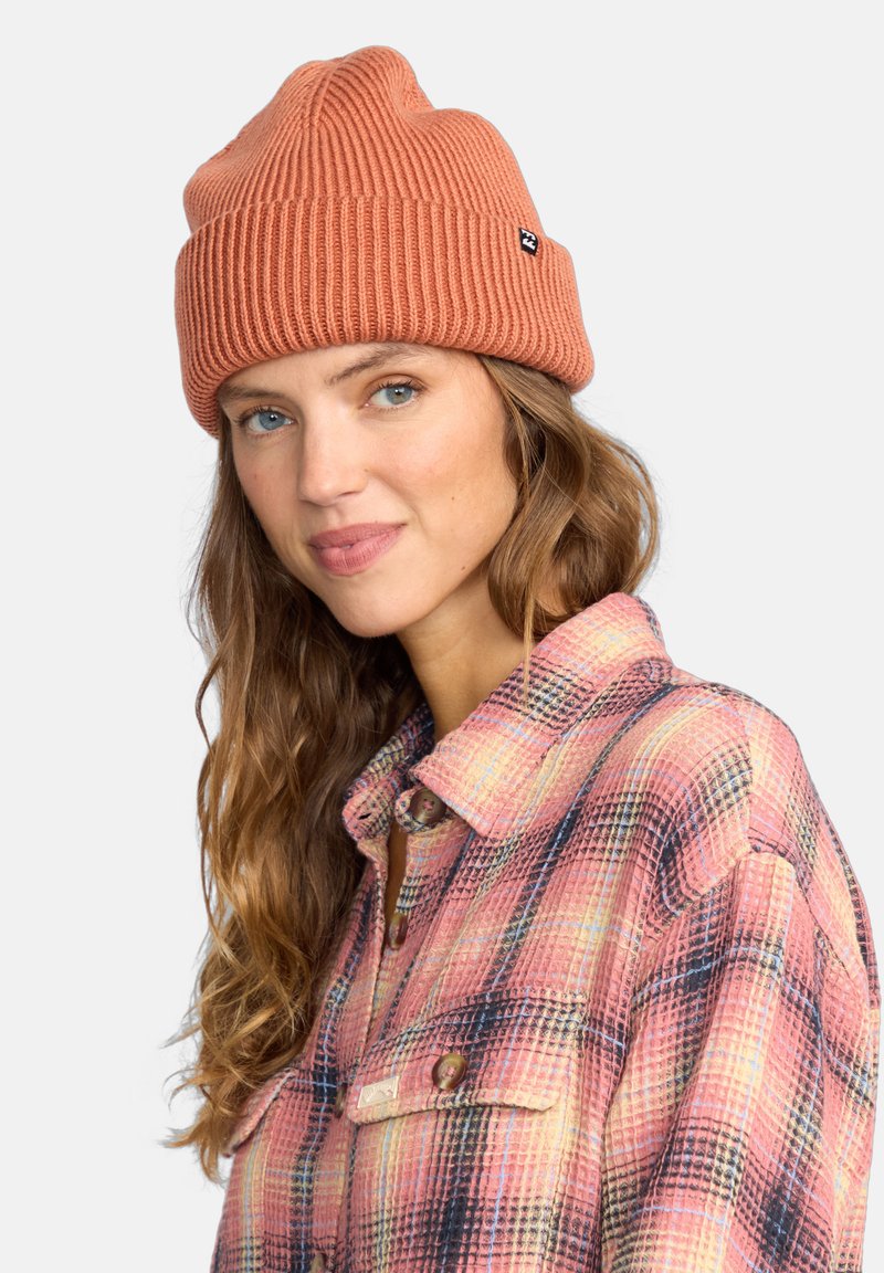 Ceglasto-czerwony dzianinowy beanie z ryżowym splotem, dopasowany krój i mała czarna metka z logo. W połączeniu z koszulą w kratę w odcieniach różu i niebieskiego.