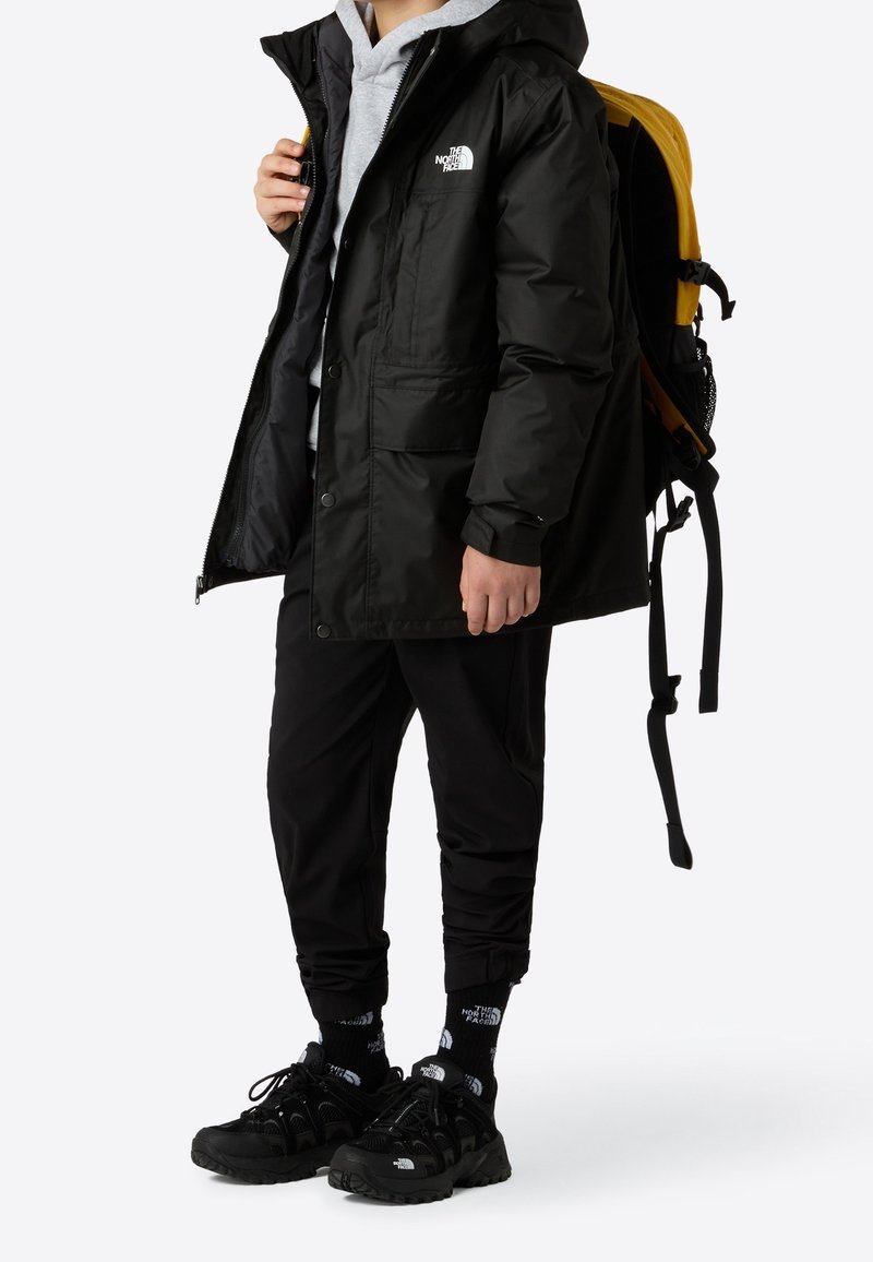 Personne portant une veste North Face noire, un pantalon noir, des chaussures noires, des chaussettes noires, un sweat à capuche gris et un sac à dos jaune et noir.