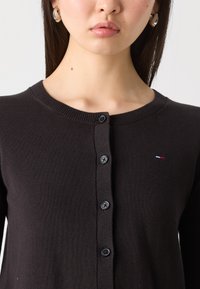 Črni pleteni kardigan z okroglim izrezom, zapiranjem na gumbe in majhnim vezenim logotipom v rdeči, beli in modri barvi na levi strani.