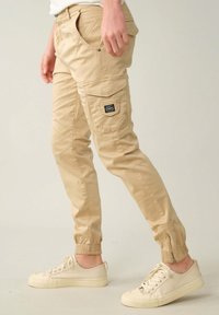 Beige cargobroek met elastische boorden, meerdere zakken, zijritsen en een stoffen label op de dij. Gemaakt van lichtgewicht materiaal.