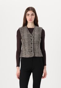 Brūns tvīda bezvīļu veste ar piestiprinātu jostu, V veida kaklu un sešiem priekšējiem pogām, saskaņota ar tumšu ilgstoša apģērba augšdaļu un melnām biksēm.