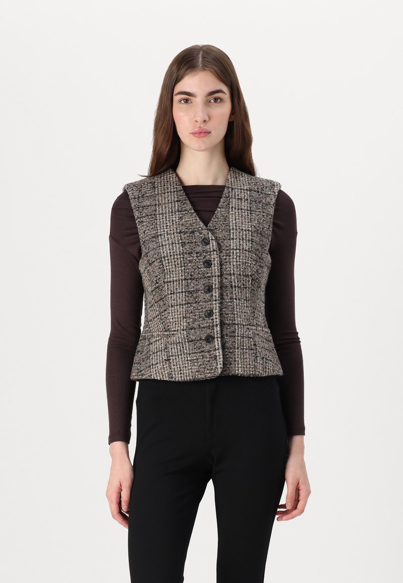 Brūns tvīda bezvīļu veste ar piestiprinātu jostu, V veida kaklu un sešiem priekšējiem pogām, saskaņota ar tumšu ilgstoša apģērba augšdaļu un melnām biksēm.