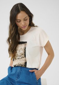 Chemise à manches courtes de couleur crème avec un graphique noir et beige portant l'inscription "LA NATURE EST UN ART." Dotée d'une coupe décontractée et d'une texture en coton douce.
