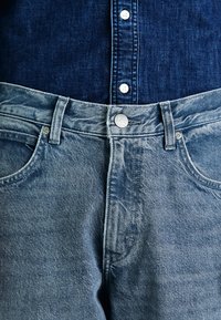 Heledad sinine denim, millel on kulunud viimistlus. Omab ümmargust halli nuppu ja nelja taskut, sileda kangast tekstuuriga.