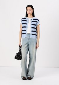Texturiertes hellblaues Strickwesten mit navyblauen Streifen und Knöpfen, kombiniert mit einem weißen T-Shirt und weit geschnittenen verwaschenen Jeans. Schwarze Taschenaccessoire.