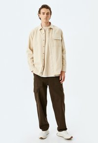 Camisa beige de botones con bolsillo en el pecho, llevada sobre una camiseta crema, combinada con pantalones cargo marrón oscuro y zapatillas blancas.