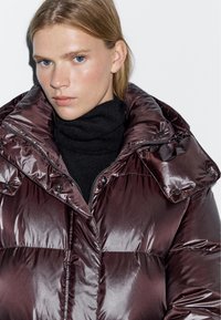 Burgundy puffer jacket with a high collar and large buttons, featuring glossy, quilted texture and a zippered front, layered over a black turtleneck.