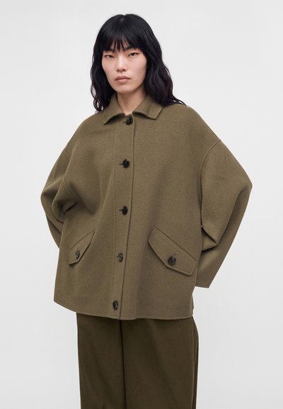 Mujer con cabello liso y oscuro que lleva un abrigo verde oliva de gran tamaño con botones, mangas anchas y bolsillos con solapa.