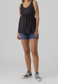 Schwarzes ärmelloses Oberteil mit drei Knöpfen und Taillenzug, kombiniert mit Jeansshorts. Das Model trägt blaue Sneakers mit weißen Schnürsenkeln.