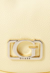Sac en cuir crème avec un logo "GUESS" argenté bien en vue. La surface présente une finition texturée avec des détails de couture subtils.
