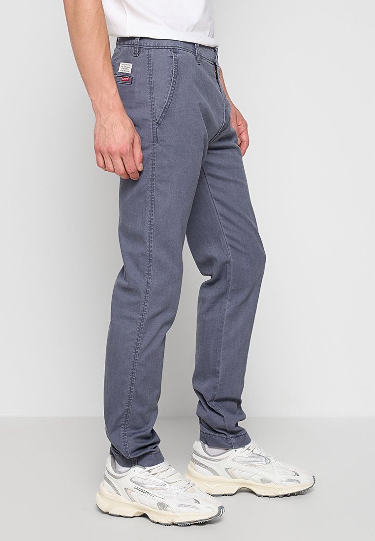 Pantalones de algodón grises con un corte recto, bolsillos frontales y una cinturilla cosida, combinados con zapatillas blancas y grises.