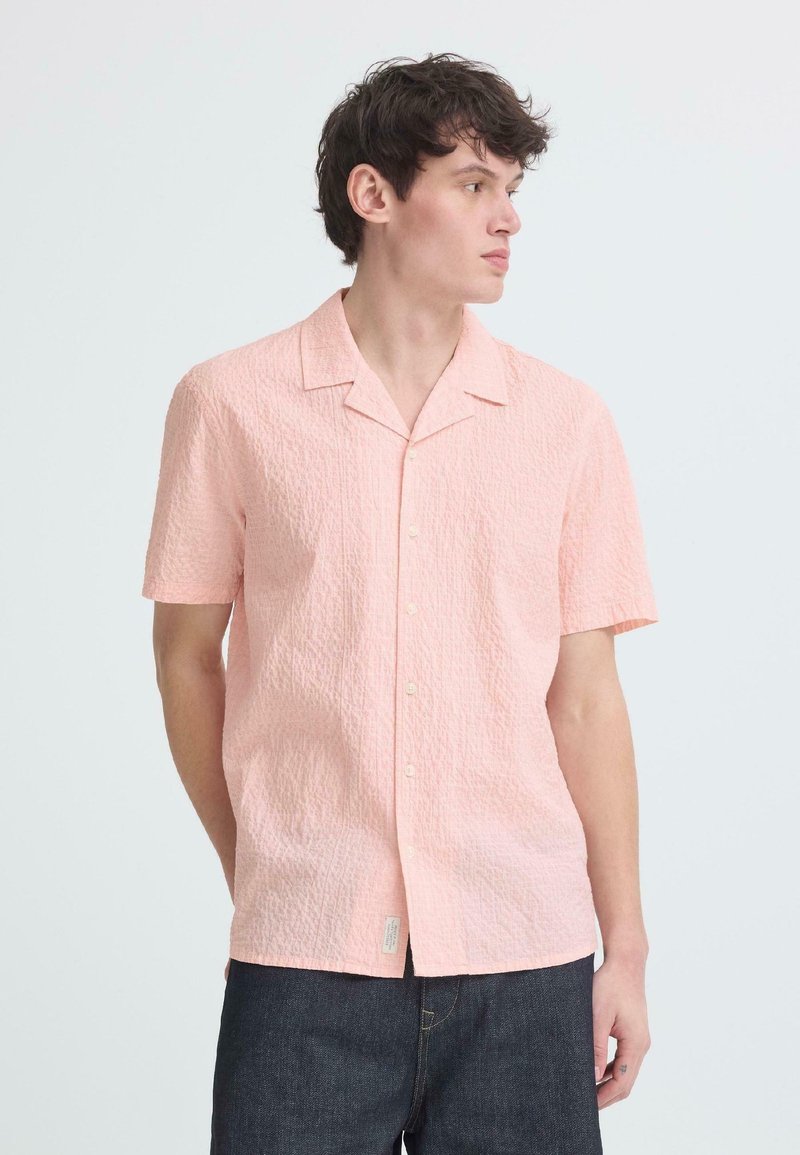 Νεαρός άνδρας που φοράει ανοιχτό ροζ κοντομάνικο πουκάμισο με υφή και σκουρόχρωμο τζιν, κοιτάζοντας προς τα δεξιά, πάνω σε απλό φόντο.