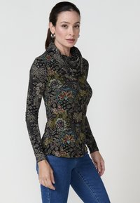Schwarz mit mehrfarbigen Blumenmustern, Oberteil mit Kapuze; lange Ärmel; figurbetontes Design; weicher, texturierter Stoff. Kombiniert mit blauen Jeans.