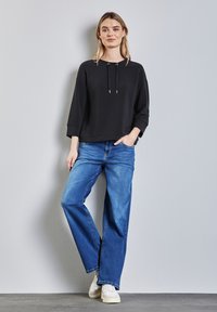 Svart sweatshirt med huva och dragskor, kombinerad med ljusblå vida jeans och vita sneakers. Enkelt, avslappnat snitt.