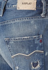 Modré džíny z denimu mají zdrsněnou texturu, charakteristické prošívání na kapsách a kožený štítek se nýty na zadní straně.