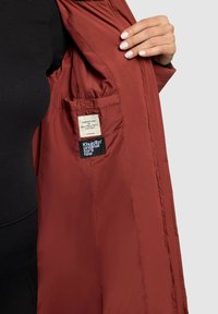 Vyno raudonos spalvos šiltai izoliuota striukė su lygiu paviršiumi, vidinėje pusėje yra kišenė ir siūti etiketės, rodanti šiuolaikišką dizainą.