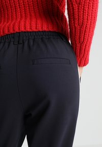 Pantalon bleu foncé avec une ceinture élastique, doté de deux poches arrière. La texture est lisse, complétée par un pull rouge côtelé.