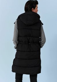 Sort polstret ærmeløs vest med hætte, der har en snøre i taljen, kontrasteret af en grå langærmet skjorte under.