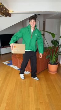 Zelená nadýchaná bunda s červeným zipsom a logom, čierne nohavice a strieborné topánky. Drží kartónovú krabicu s nápisom „Miluj ma. Noste ma.“