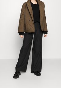 Prošívaná hnědá bunda s velkým límcem a dvojitým zapínáním na knoflíky, doplněná černými širokými kalhotami a robustními černými botami.