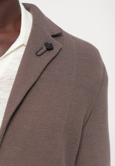 Brown knit blazer with textured material, featuring a black floral lapel pin and subtle notched collar. Layered over a white shirt.
