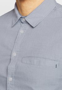 Vaaleansininen kauluspaita, jossa on terävä kaulus ja pieni tasku vasemmassa rinnassa, päällä henkilö, jonka kaula ja leukalinja ovat näkyvissä.